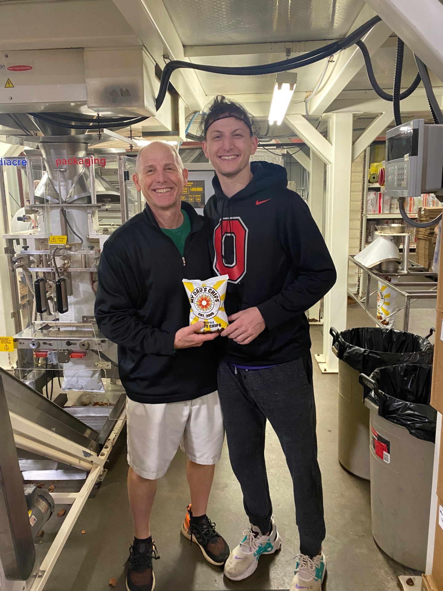 Gary and Brian holding a bag of chips beside the packaging line at the production facility.